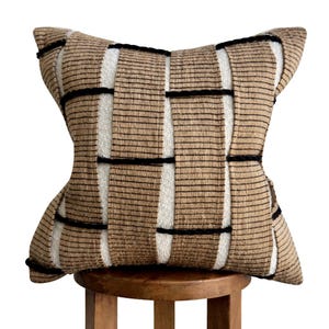 May include: Square cushion with a woven design. The cushion has a tan background with black lines and white stripes. The cushion is placed on a wooden stool. The cushion measures approximately 40cm x 40cm.