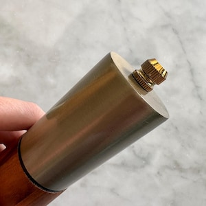 May include: A close-up of a silver and wood pepper mill with a gold top. The mill is held in a hand.