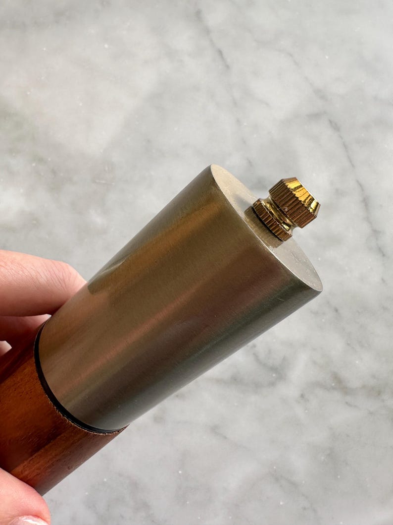 May include: A close-up of a silver and wood pepper mill with a gold top. The mill is held in a hand.