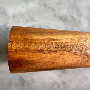 May include: A close-up of a wooden rolling pin with a smooth, brown surface. The rolling pin is made of natural wood and has a slightly rounded shape.