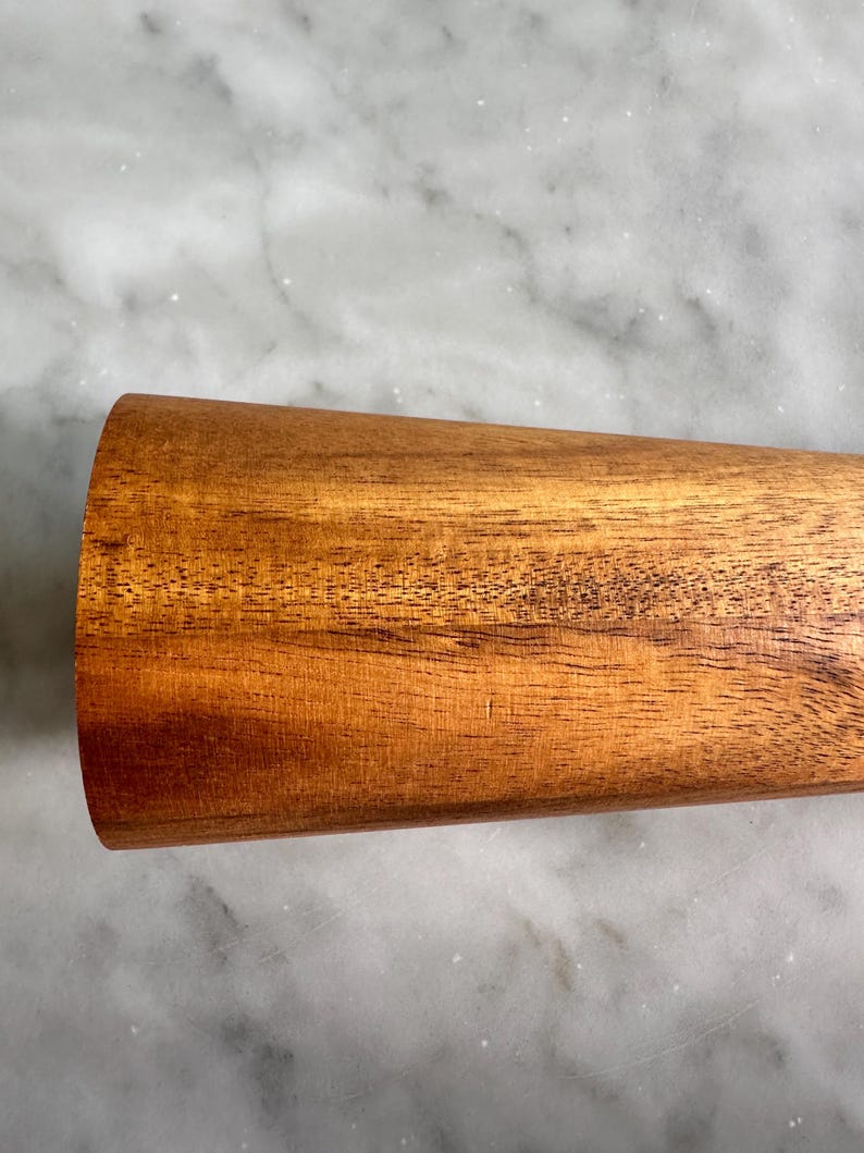 May include: A close-up of a wooden rolling pin with a smooth, brown surface. The rolling pin is made of natural wood and has a slightly rounded shape.
