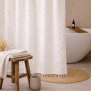 May include: White shower curtain with a textured chevron pattern and tassel trim. A wooden stool holds folded towels. A round woven rug and a stone-colored container are on the floor. A bathtub is in the background.