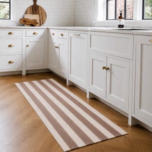May include: A rectangular runner rug featuring alternating vertical stripes in muted brown and cream tones. The rug is positioned on a light brown wooden floor in a kitchen with white cabinetry, brass hardware, and a white brick backsplash.