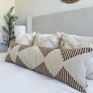 Peut inclure: Un long coussin rectangulaire avec un fond crème et un motif répété de triangles bruns pointant vers le bas. Le coussin est placé sur un lit blanc avec d'autres coussins décoratifs. Le lit a une tête de lit gris clair.