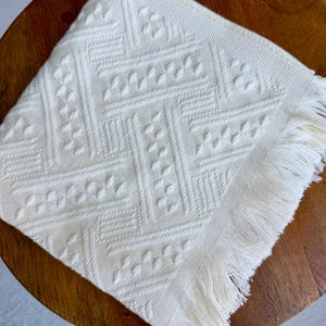 May include: Ivory-colored square cloth napkin with a geometric pattern and fringed edge. The napkin is placed on a wooden surface. The pattern includes a series of diamond and floral shapes. The napkin is made of a soft, textured fabric.