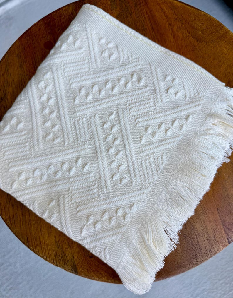 May include: Ivory-colored square cloth napkin with a geometric pattern and fringed edge. The napkin is placed on a wooden surface. The pattern includes a series of diamond and floral shapes. The napkin is made of a soft, textured fabric.