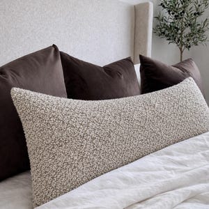 Puede incluir: Un cojín lumbar largo y rectangular con una funda de tela bouclé texturizada de color crema. El cojín está colocado horizontalmente frente a tres cojines cuadrados de color marrón oscuro y superficie lisa, dispuestos contra un cabecero claro.