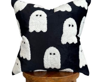May include: Black velvet pillow with white fluffy ghost appliques. The pillow is sitting on a wooden stool.
