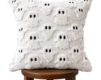 May include: A square white throw pillow featuring a repeating pattern of raised, textured ghost figures. Each ghost is rendered in white plush fabric with two small, embroidered black circular eyes, creating a consistent, tactile design across the surface.