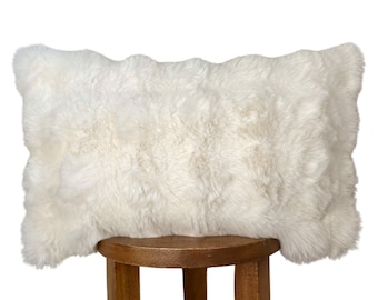 Puede incluir: Un cojín de piel de oveja blanca con textura esponjosa. El cojín está sentado en un taburete de madera.