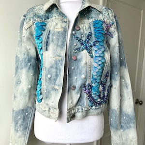 May include: A light blue denim jacket with pearl embellishments and a mermaid-themed design featuring blue and purple sequins, seashells, and a starfish.