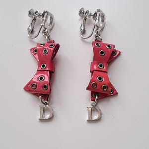Peut inclure: Une paire de boucles d'oreilles clips en forme de nœud rouge avec des accents en métal argenté et un charme 'D' argenté suspendu à chaque nœud.
