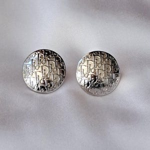 May include: A pair of silver-coloured, circular earrings with a repeating pattern. The earrings are set against a white, silky backdrop. The pattern appears to be a repeating logo or design. The earrings are reflective and shiny.