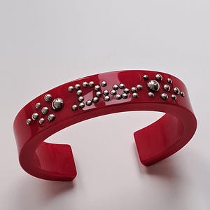 Könnte beinhalten: Ein rotes Plastik-Armband mit silbernen Nieten, die in einer stilisierten Schrift "Dior" buchstabieren.