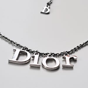May include: Silver-toned necklace featuring the letters "DIOR" as pendants. The necklace has a delicate chain and a small "D" charm near the clasp. The letters are in a bold, block style, creating a modern and elegant look.
