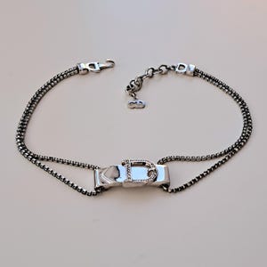 May include: A silver-toned necklace featuring a buckle design with a 'D' detail and sparkling accents. The necklace has a double-strand chain and a hook clasp. The overall design is elegant and suitable for formal occasions.