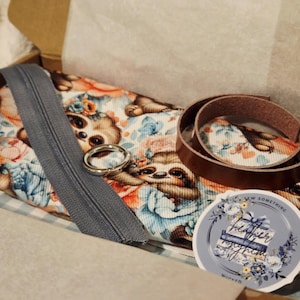 May include: A craft kit in a cardboard box, featuring a sloth-themed fabric, a gray zipper, a silver ring, and a brown leather strap. A sticker with floral accents and text is also included. The fabric has a blue and orange floral pattern.