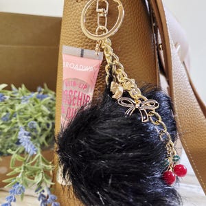 May include: A gold-toned keychain with a black fluffy pom-pom, a small tube of lip gloss, and a decorative chain with a bow and red cherry-like beads. The keychain is attached to a brown leather handbag.