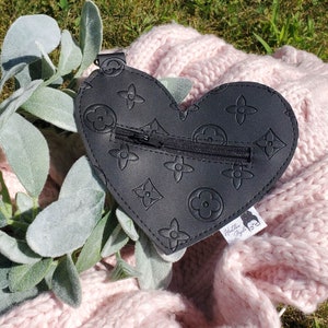 Puede incluir: Bolsa negra con cremallera en forma de corazón con un patrón de flores en relieve repetido. La bolsa tiene una etiqueta blanca con el texto "Wilder My Style".