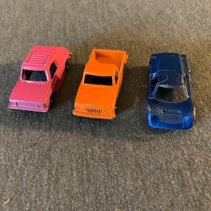 May include: Three toy cars in bright colors: a pink SUV, an orange pickup truck, and a blue sports car. The vehicles are made of metal and have detailed features like windows and grilles. The cars are arranged on a gray surface.