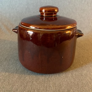 May include: A brown ceramic casserole dish with a lid. The dish has a round shape with two small handles on the sides. The lid has a small knob on top for easy handling. The dish is glazed with a glossy finish.
