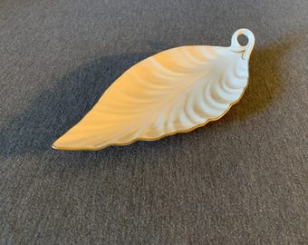 May include: A leaf-shaped decorative dish with a cream and gold color scheme. The dish has a gold trim and a small loop at the end. The design features a pattern of curved lines, resembling the veins of a leaf. This item is suitable for serving small items.