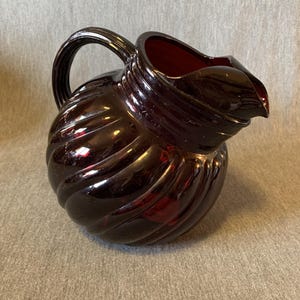 Puede incluir: Una jarra de vidrio rojo oscuro con un diseño en espiral. La jarra tiene un asa y un pico para verter. El vidrio es brillante y refleja la luz, dándole un aspecto vintage. La jarra probablemente se utiliza para servir bebidas.