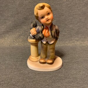 Puede incluir: Figura de porcelana de un niño en traje, sosteniendo un objeto negro sobre un pedestal. El niño tiene el pelo rubio y lleva un chaleco blanco, pantalones marrones y una corbata roja. La figura está sobre una base redonda blanquecina.