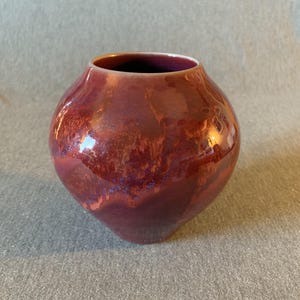 May include: A round, ceramic vase with a glossy, reddish-brown glaze. The vase has a wide body and a narrow opening. The glaze has a mottled appearance with hints of purple and blue, creating a unique, decorative piece.