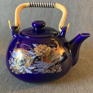 Pode incluir: Uma chaleira de cerâmica azul cobalto com alça e tampa de bambu. A chaleira é decorada com desenhos dourados e brancos de pavão e flores. A alça é bege claro com listras azul escuro. O bico está no lado direito.