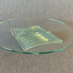 Può includere: Un piatto di vetro trasparente con un centrotavola decorativo in vetro geometrico. Il centrotavola è composto da diversi strati di vetro curvo, creando un effetto a strati. Il piatto è rotondo e ha un bordo liscio e lucido.