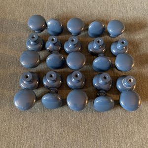 May include: A collection of twenty round, glossy, slate blue cabinet knobs arranged in four rows of five. Each knob features a smooth, domed top and a central metal-lined screw hole on the underside for attachment to furniture or cabinetry.