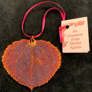 May include: A gold leaf ornament shaped like a leaf with a pink ribbon attached. The ornament has a tag that reads "An Ornament From Mother Nature".