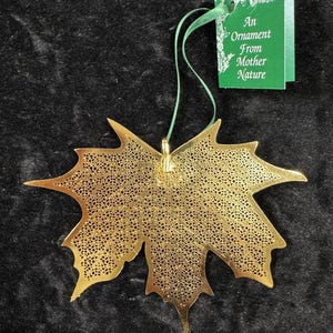 May include: A gold-colored, leaf-shaped ornament with intricate cut-out details. The ornament is suspended by a green ribbon and has a green tag that reads "An Ornament From Mother Nature".