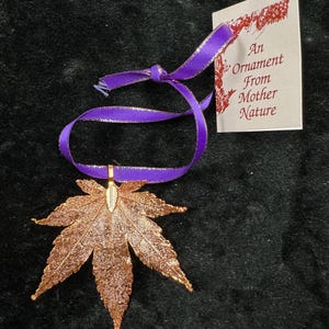 May include: A copper-colored leaf ornament with a purple ribbon. The leaf has intricate details and is attached to a small gold-colored loop. A tag with the text "An Ornament From Mother Nature" is attached to the ribbon.