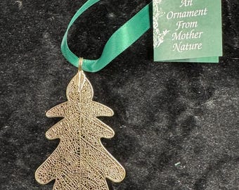 May include: A gold-colored leaf ornament with intricate detailing, suspended by a green ribbon. A green tag reads "An Ornament From Mother Nature". The ornament is set against a dark, textured background.