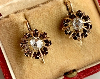 May include: A pair of antique gold earrings with a floral design. Each earring features a central, faceted, clear gemstone surrounded by gold petals and dark accents. The earrings are displayed in a red velvet jewelry box.