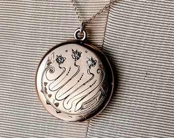 May include: A round, rose gold locket pendant with an ornate floral design and three small, sparkling gemstones. The pendant hangs from a delicate silver chain against a striped background.