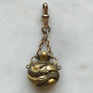 May include: A vintage gold-toned pendant with a small, round perfume bottle design. The bottle features floral and beaded accents, with a chain and clasp for attachment. The pendant is likely made of brass or a similar metal.