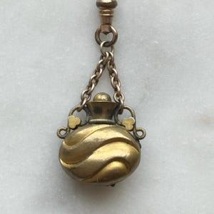 May include: Antique gold-toned pendant with a small, round perfume bottle design. The bottle features a swirling pattern and is suspended from a chain and clasp. Decorative heart-shaped accents adorn the sides of the bottle, adding a vintage charm.