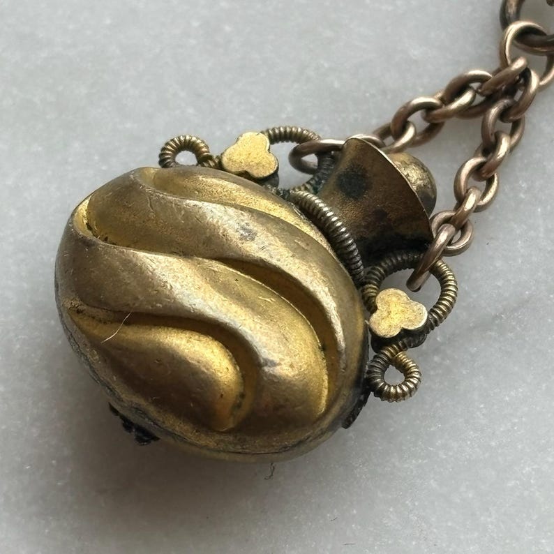 May include: A vintage gold-toned pendant necklace. The pendant is an oval shape with a textured, swirling design. It has decorative floral accents and is attached to a delicate chain. The metal has an aged appearance.
