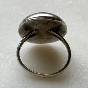 May include: A silver ring with a round, smooth, silver top. The ring is made of a single band of metal.