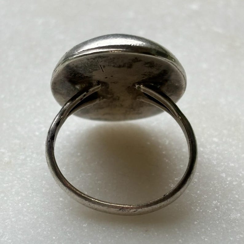 May include: A silver ring with a round, smooth, silver top. The ring is made of a single band of metal.