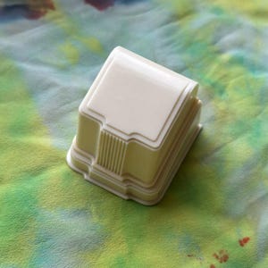 Pode incluir: Uma caixa de anel de plástico branco com um design decorativo. A caixa tem uma tampa elevada e uma base com um design estriado.