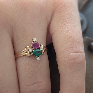 Könnte beinhalten: Ein goldener Ring mit einem Rubin- und Smaragd-Edelstein-Design, akzentuiert mit kleinen Diamanten. Der Ring hat eine verzierte, von Vintage inspirierte Fassung mit detaillierten Filigranarbeiten. Die Edelsteine sind oval geschliffen und in einer Cluster-Anordnung angeordnet.