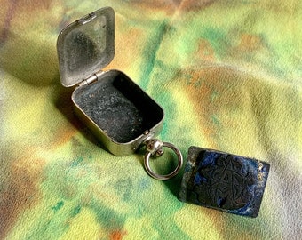 May include: A silver metal box with a hinged lid, open to reveal an empty interior. The box is attached to a silver ring and a black square stamp with a decorative design.