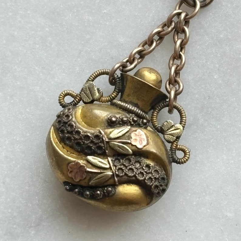 May include: An antique gold-toned perfume bottle pendant with a decorative chain. The bottle features intricate detailing, including floral accents and a textured design. The pendant is a vintage piece, showcasing a classic aesthetic.