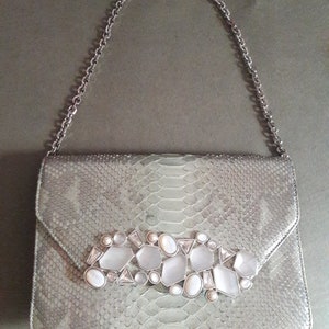 May include: A silver chain strap clutch purse with a snakeskin pattern and a large silver and white rhinestone embellishment.