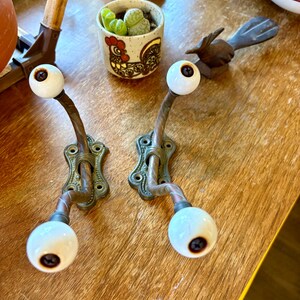 May include: Two antique brass coat hooks with white ceramic knobs and dark brown centers. The hooks have ornate, decorative backplates. A small ceramic pot with a rooster design and a small wooden bird are also visible.