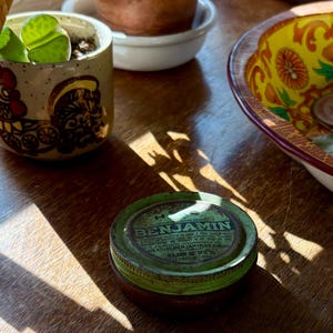 May include: A still-life composition featuring a small, decorative ceramic pot with a rooster design, a vintage green tin labeled "BENJAMIN", and a colorful bowl with floral patterns. The items are arranged on a wooden surface, illuminated by sunlight.